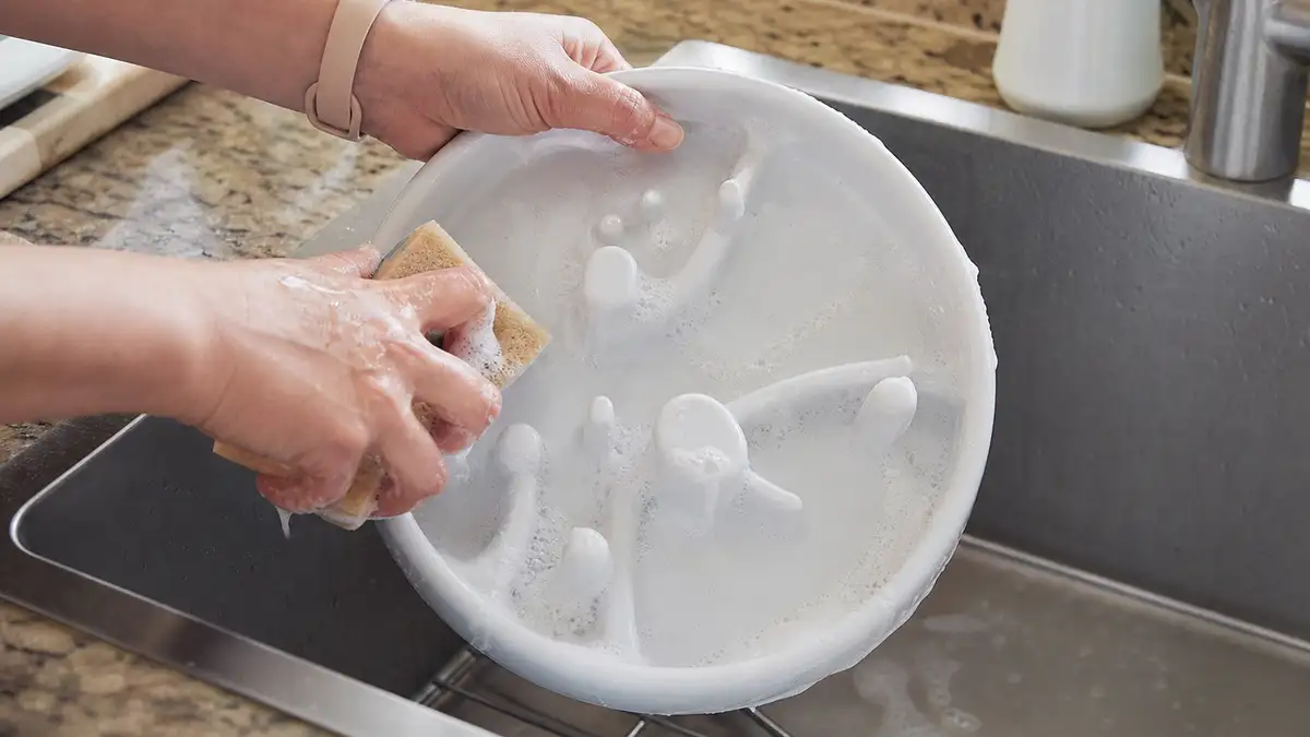 Hands washing a round plastic pet feeder food tray in a kitchen sink